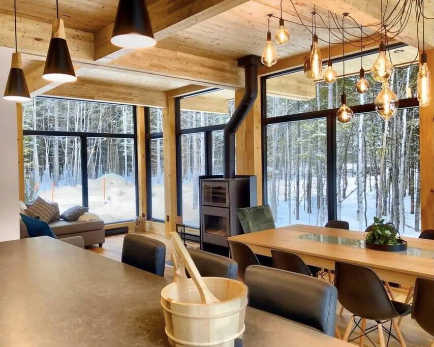 An open-plan living area features large glass windows offering views of the snowy exterior. A wooden dining table accommodates guests, surrounded by black chairs. A modern fireplace and a stylish lighting fixture with hanging bulbs contribute to the space's design. Warm wooden accents add to the inviting ambiance.