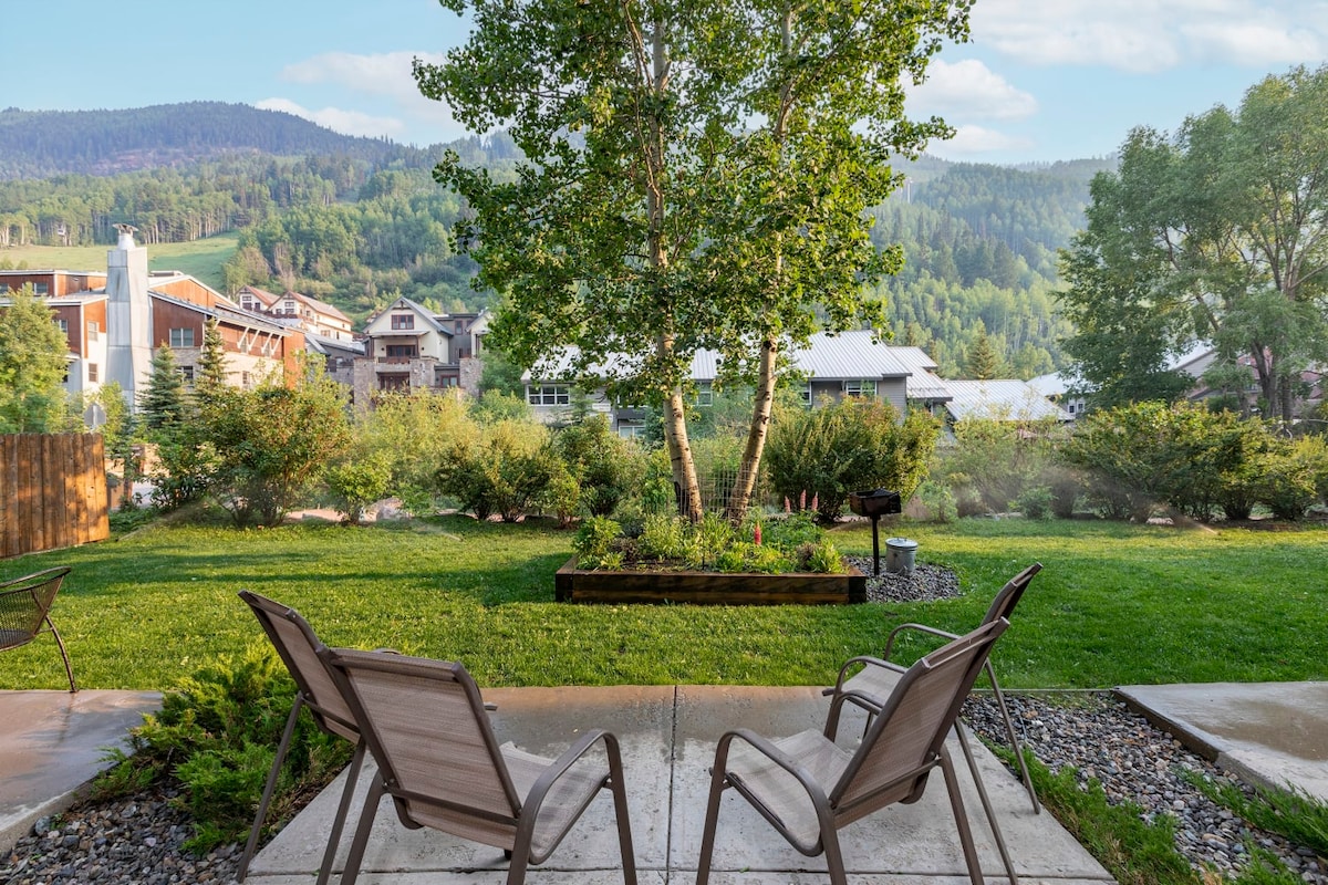 Two patio chairs are positioned on a stone surface, facing a lush green lawn and trees. A raised garden bed sits in the foreground, surrounded by low vegetation. In the background, a scenic view of rolling hills and buildings is visible under a clear sky.