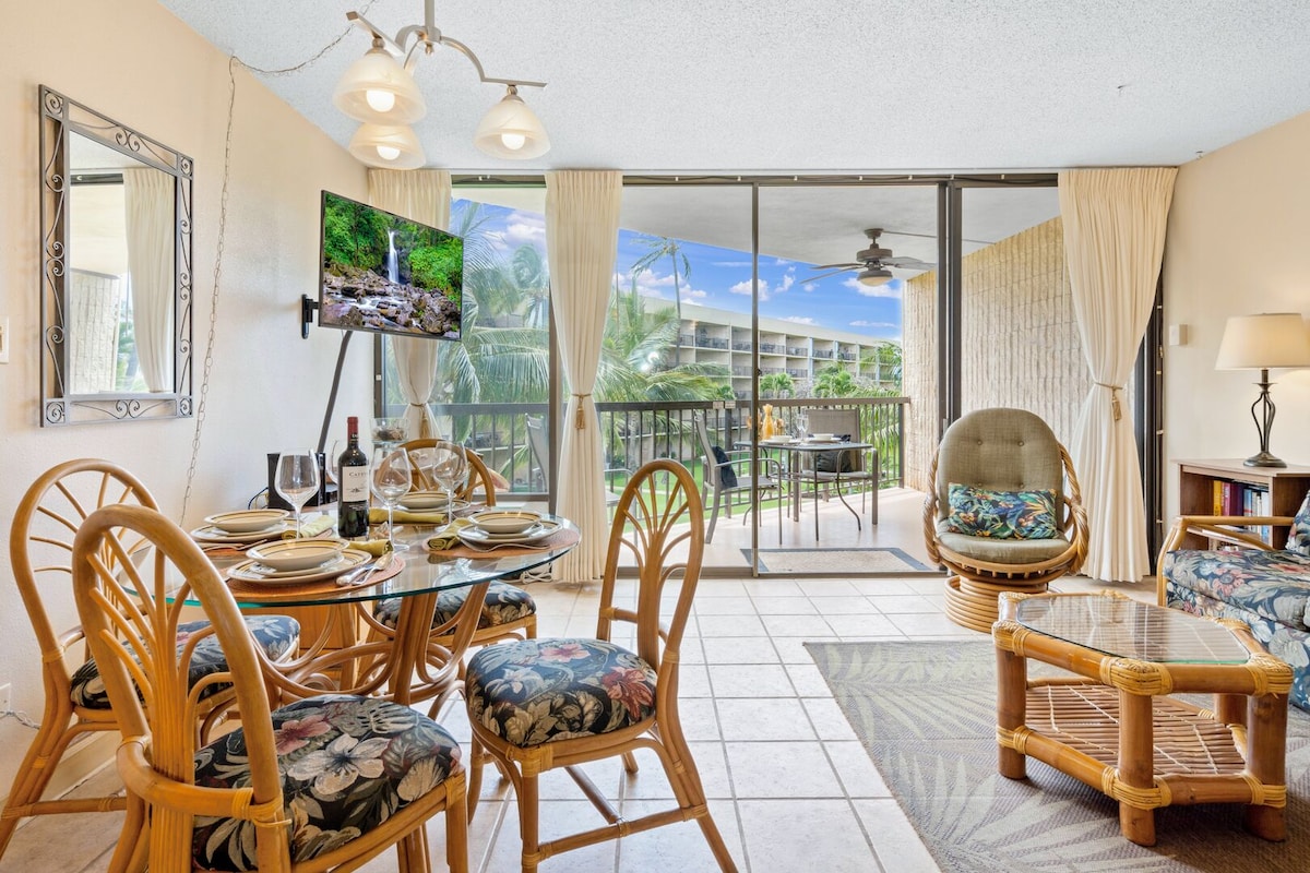 A spacious living area is furnished with a glass dining table set for four, accompanied by comfortable seating. Large sliding doors open to a private balcony, allowing natural light to fill the room. Tropical decor enhances the space, creating a relaxing environment.