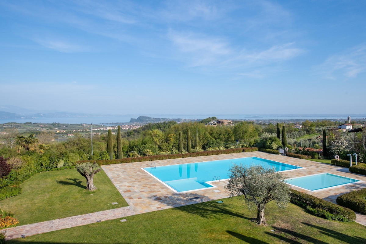 A tranquil landscape features a large swimming pool surrounded by well-maintained gardens. Olive trees and neatly trimmed hedges frame the area, while panoramic views of Lake Garda and distant hills stretch beyond. Clear blue skies and soft clouds complete the serene atmosphere.