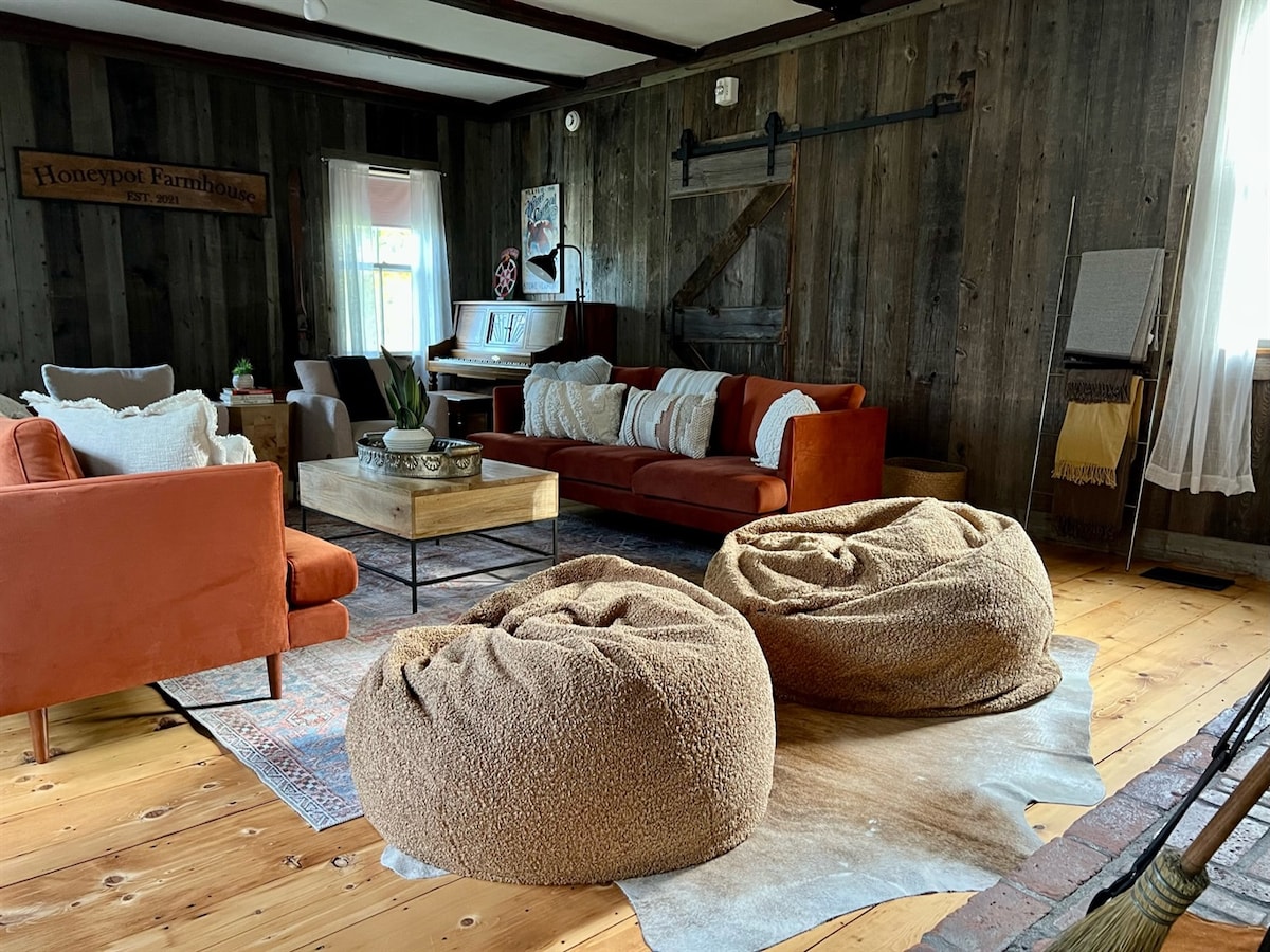A cozy living space features soft bean bag chairs in earth tones, accompanied by a rustic coffee table and a comfortable seating area with orange and cream sofas. The wood-paneled walls and light streaming through sheer curtains add warmth to the environment.