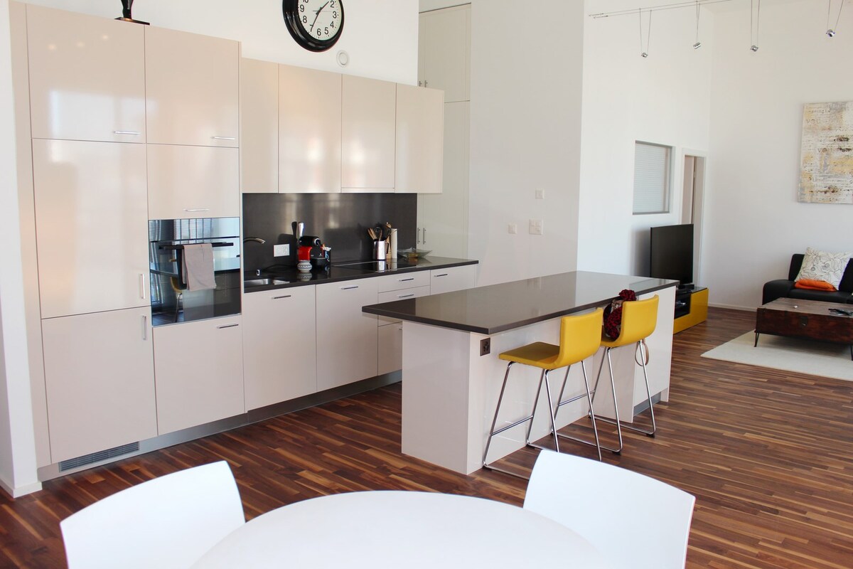 A modern kitchen space features sleek cabinetry and a dark countertop. Bar stools with yellow seats are positioned at the island, while a circular dining table with white chairs is visible. A clock hangs on the wall, complementing the minimalist aesthetic.