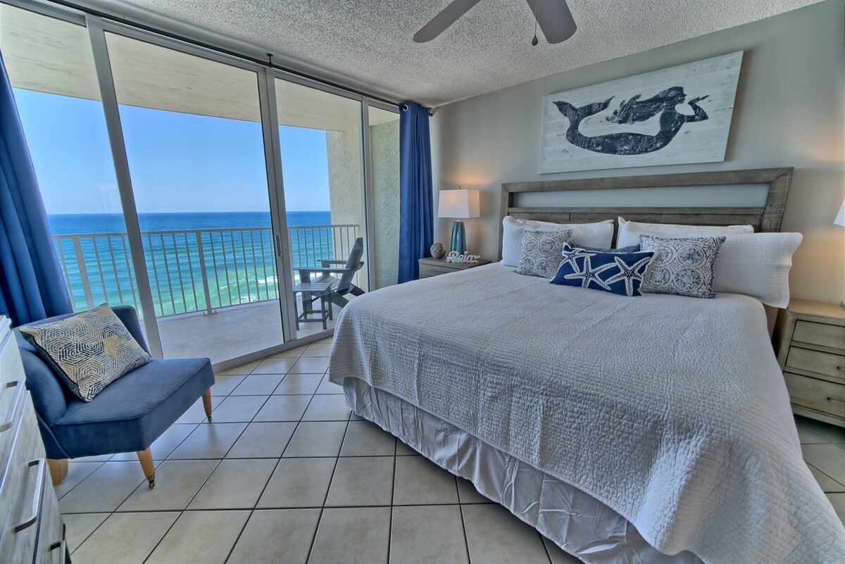 A spacious bedroom features a king bed dressed in white linens, complemented by decorative pillows. A blue accent chair is positioned beside the bed. Large glass doors offer expansive views of the Gulf of Mexico, allowing natural light to fill the room.