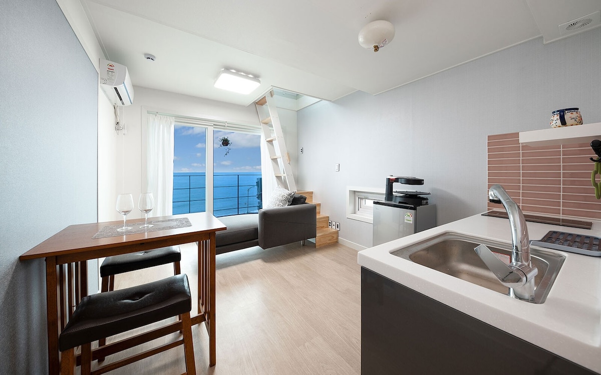 A bright living space features a modern kitchenette, a dining table with two chairs, and a cozy seating area with a sofa. Large windows provide unobstructed ocean views, while a staircase leads to an upper lofted space.