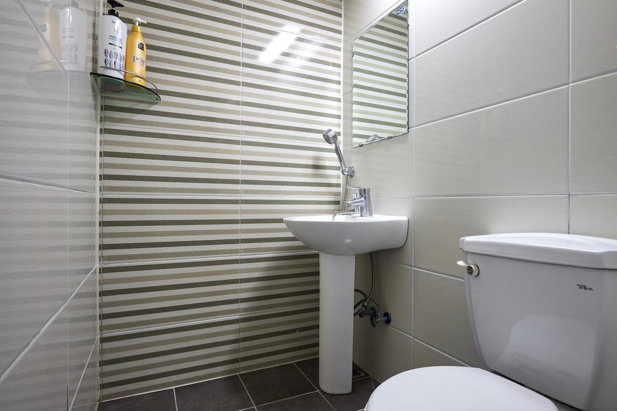 A compact bathroom features tiled walls with a striped pattern. A wall-mounted shelf holds toiletries. A white sink and a modern toilet are visible, with natural light reflected off the mirror, enhancing the space's cleanliness and functionality.