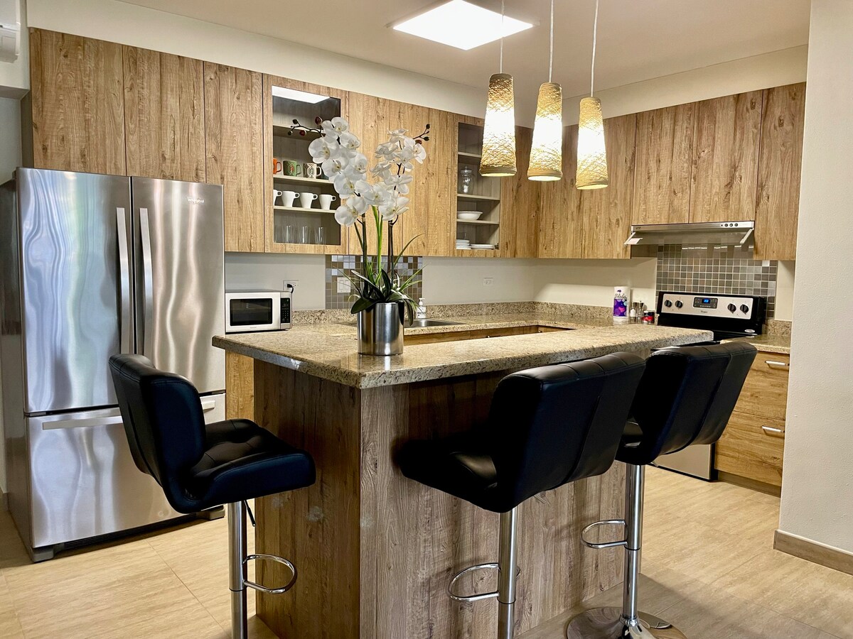 The modern kitchen is equipped with sleek stainless-steel appliances and wooden cabinetry. A spacious kitchen island with seating for four contrasts with the light-colored tile flooring. Decorative elements include a vase of white orchids and pendant lighting that adds a warm ambiance to the space.