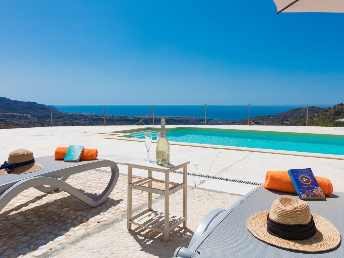 Two deck chairs are positioned by a swimming pool, with a small table holding glasses and a bottle. Sun hats rest atop the chairs. A clear blue sky and distant sea views create a serene backdrop for relaxation.