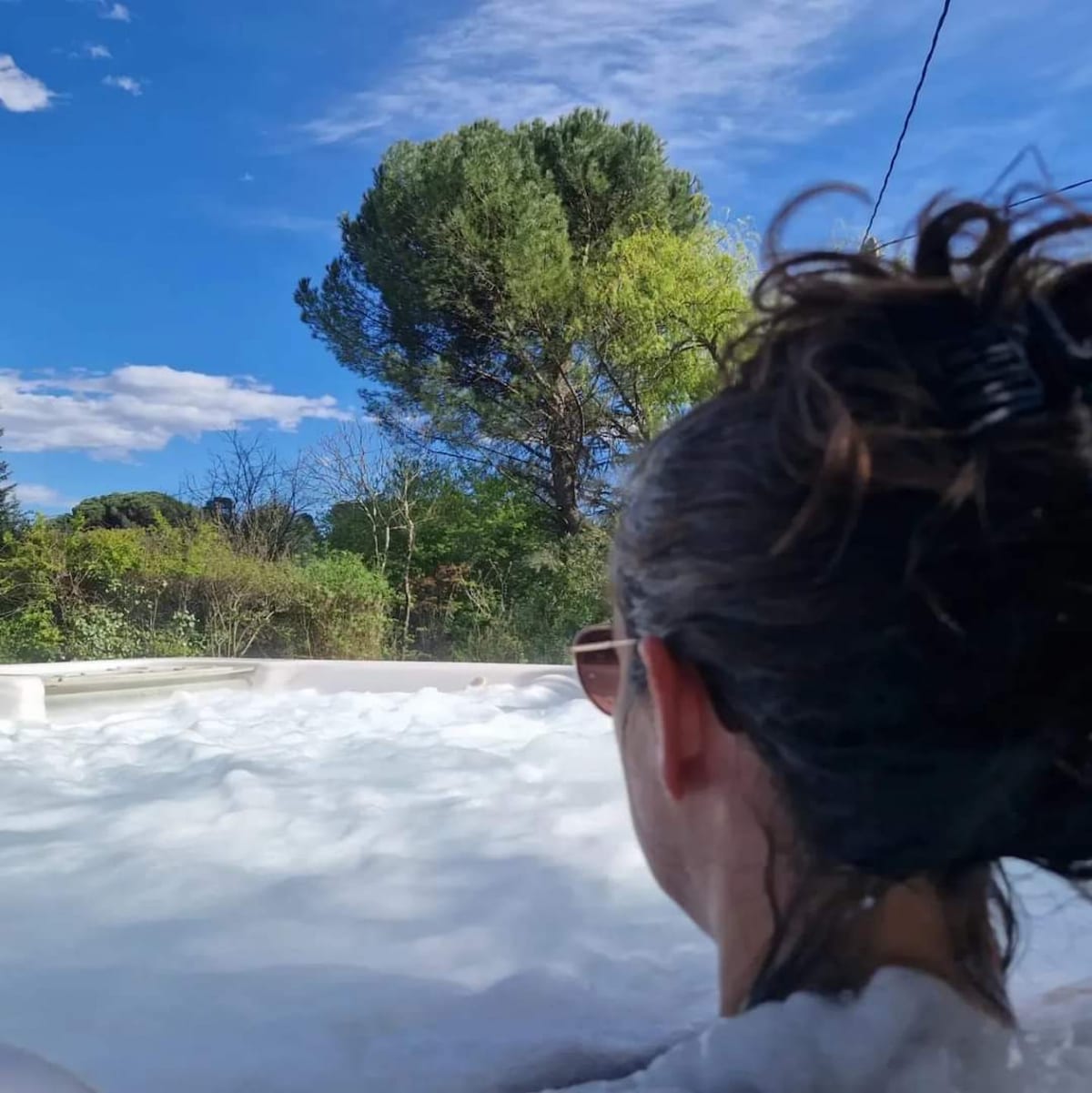A bubbling jacuzzi is shown under a clear blue sky dotted with clouds. In the foreground, a person with curly hair is partially visible, facing away from the camera, while lush greenery and trees surround the area, enhancing the serene atmosphere.