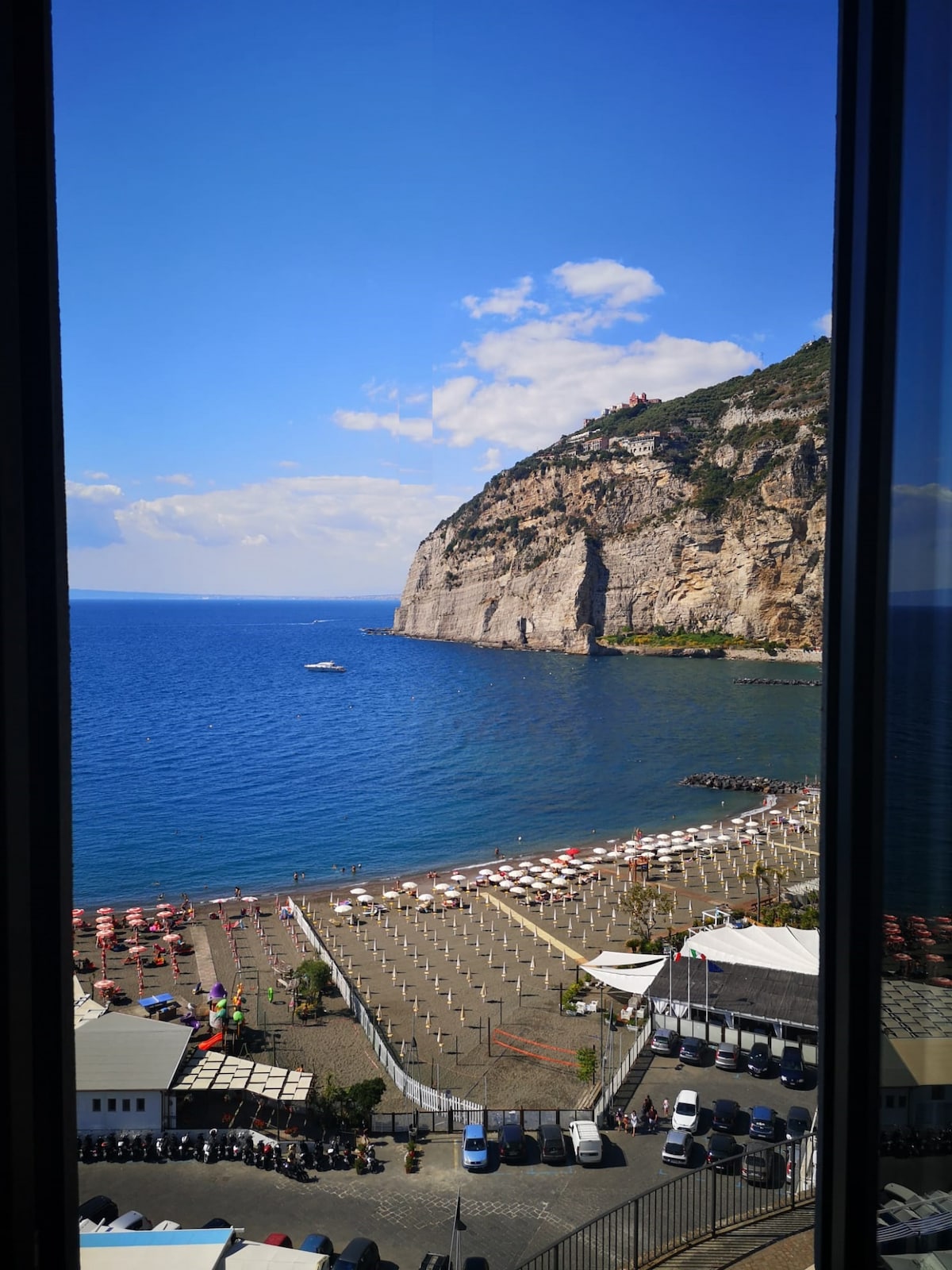 A panoramic view of the coastline is showcased through a large window. The image features a rocky cliff against a backdrop of a clear blue sky, with the sparkling waters below. The beach is lined with beach umbrellas and sunbeds, indicating a vibrant seaside atmosphere.