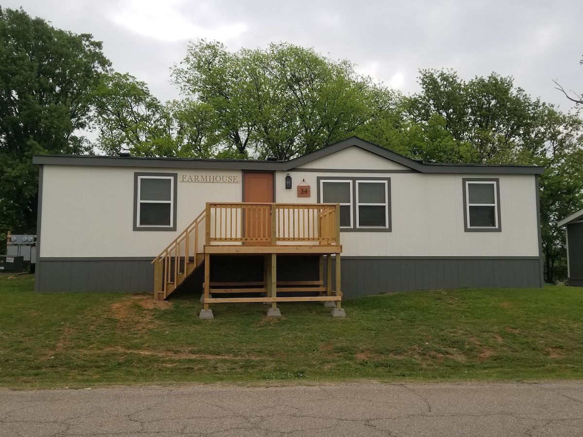 A rustic farmhouse featuring a welcoming wooden front porch accessed by a ramp. The structure has a simple two-tone exterior with large windows, surrounded by green grass and trees, providing a connection to the natural environment.