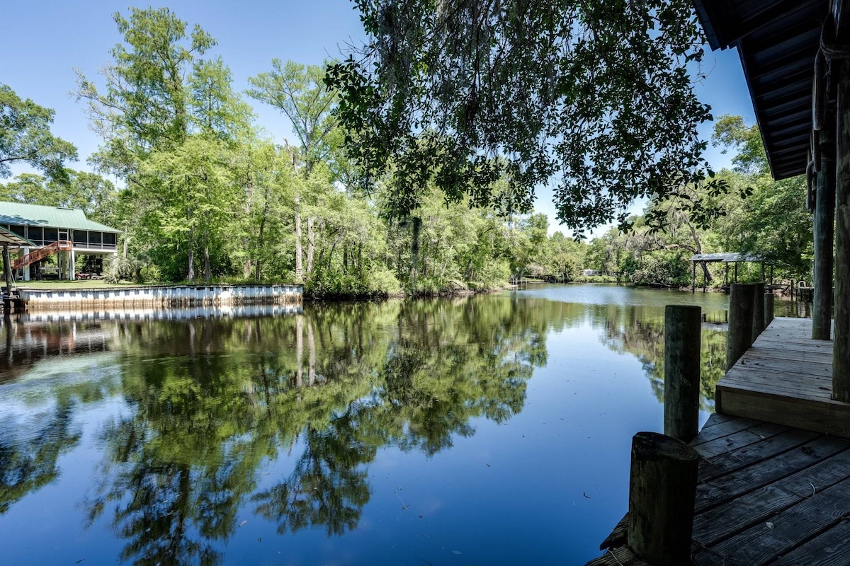The tranquil Black Creek reflects the surrounding greenery, with dense trees lining the water's edge. A wooden dock extends into the calm water, offering a serene view across the creek, where wildlife may be spotted among the lush foliage.