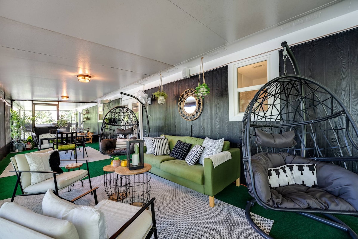 A spacious screened-in porch features a mix of seating options, including hanging chairs and a green sofa. Natural light filters through large windows, illuminating the area adorned with plants and decorative elements, creating a cozy atmosphere for relaxation.