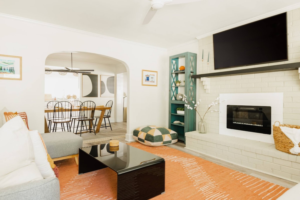 A bright living room features a comfortable gray sofa with colorful accent pillows. A black coffee table sits on a light orange area rug, while a modern fireplace and mounted TV enhance the space. A dining area is visible, furnished with a wooden table and black chairs.