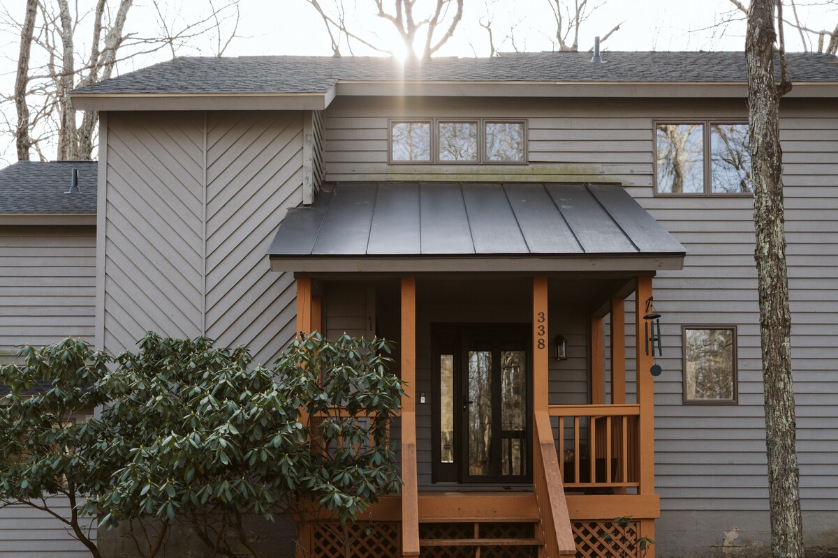 The cabin's entrance features a welcoming porch with an overhang, providing shelter. Large windows above the door allow natural light to fill the area. Surrounding greenery adds a natural touch, enhancing the sense of tranquility in this wooded setting.