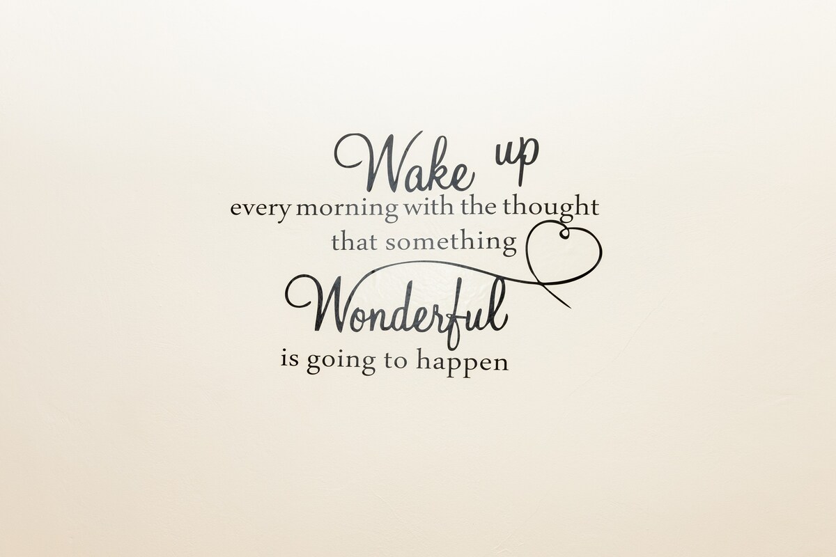 A wall is adorned with a decorative quote that reads, 'Wake up every morning with the thought that something wonderful is going to happen.' The lettering features a mix of elegant fonts, set against a soft light background, adding a stylish touch to the space.