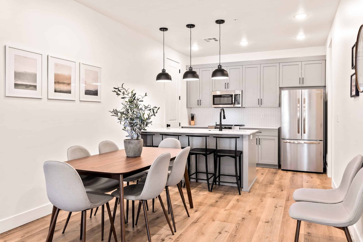 A modern dining area is featured, showcasing a wooden table surrounded by six light grey chairs. Above, three black pendant lights illuminate the space. The kitchen includes stainless steel appliances and light-grey cabinetry, creating a clean and functional setup.