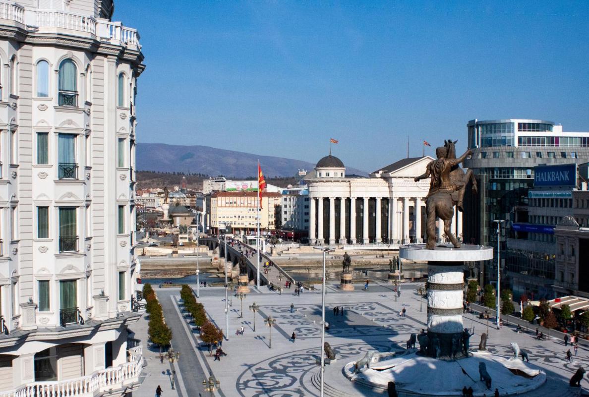 A panoramic view of Macedonia Square is depicted, featuring the statue of Alexander the Great at its center. Buildings with diverse architectural styles surround the square, and pedestrians can be seen enjoying the open space. The distant hills create a serene backdrop.