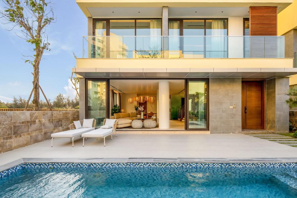A modern villa is showcased, featuring an inviting outdoor pool with a mosaic edge. Spacious sliding glass doors open to an airy living area, allowing natural light to fill the interior. Two sun loungers are positioned on the pool deck, complemented by greenery in the background.