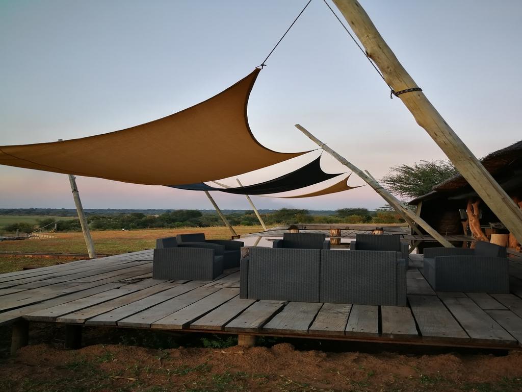 A spacious wooden deck features a seating area furnished with black wicker chairs and tables. Canvas shades provide shelter while blending with the surrounding natural landscape. The view includes distant greenery and open skies, offering a serene outdoor space.