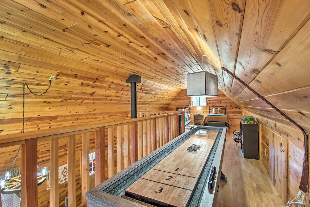 An inviting loft space features warm wooden paneling and a shuffleboard table, contributing to a cozy atmosphere. Natural light filters through windows at both ends, illuminating the area. A wooden railing overlooks the main level, while additional seating is visible in the background.