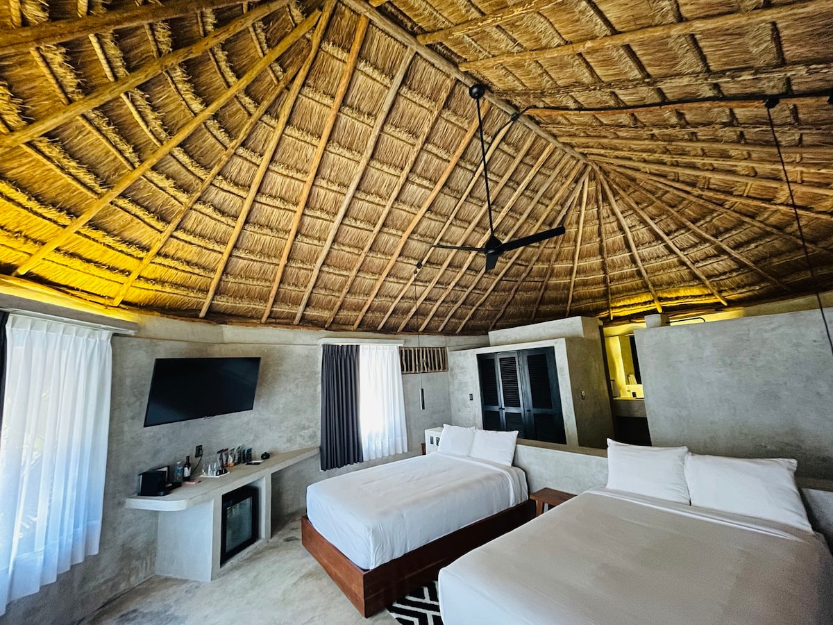 A spacious room features a unique thatched ceiling, enhancing the rustic charm. Two beds with white linens are positioned opposite a wall-mounted TV. Natural light filters through sheer and blackout curtains, while a small seating area is complemented by a mini-fridge and a coffee station.
