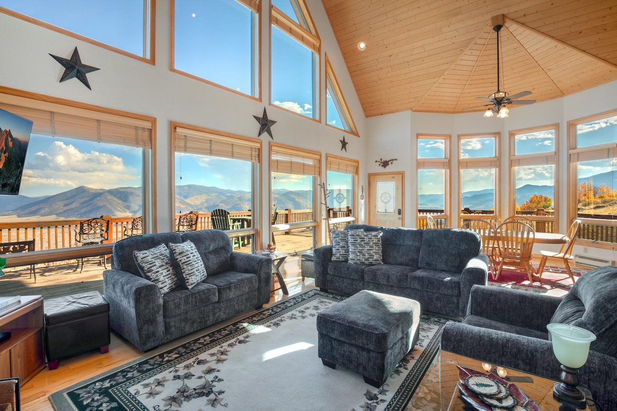 A spacious living area features large windows that provide panoramic views of the surrounding mountains. Comfortable seating is arranged around a central rug, and natural light fills the room. Sliding doors lead to an expansive outdoor deck, enhancing the connection to the scenic landscape.