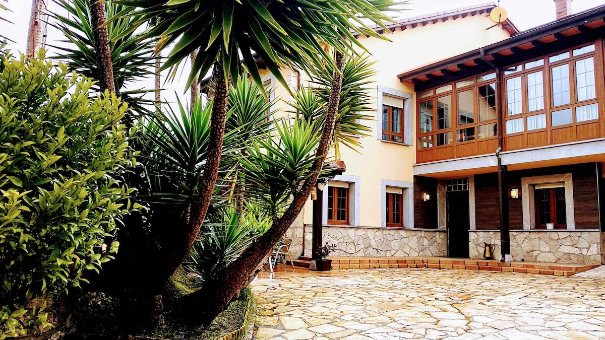 A landscaped exterior features a stone-paved patio surrounded by lush tropical plants and shrubs. The two-story building is characterized by large windows with wooden accents and a covered porch area, inviting relaxation in a natural, serene environment.