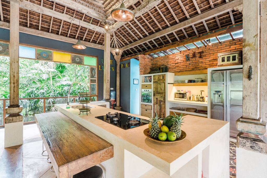 A spacious kitchen features a large wooden island with a built-in stove, surrounded by fresh fruits. Open shelving displays kitchen essentials, while modern appliances are integrated into a brick and wooden design. Large windows provide a view of the lush greenery outside.