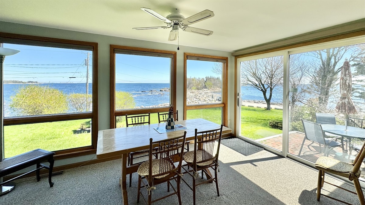 A dining area is presented with large windows offering views of the ocean and surrounding greenery. A spacious wooden table is surrounded by several chairs, and a sliding glass door leads to an outdoor area. Bright natural light fills the room.
