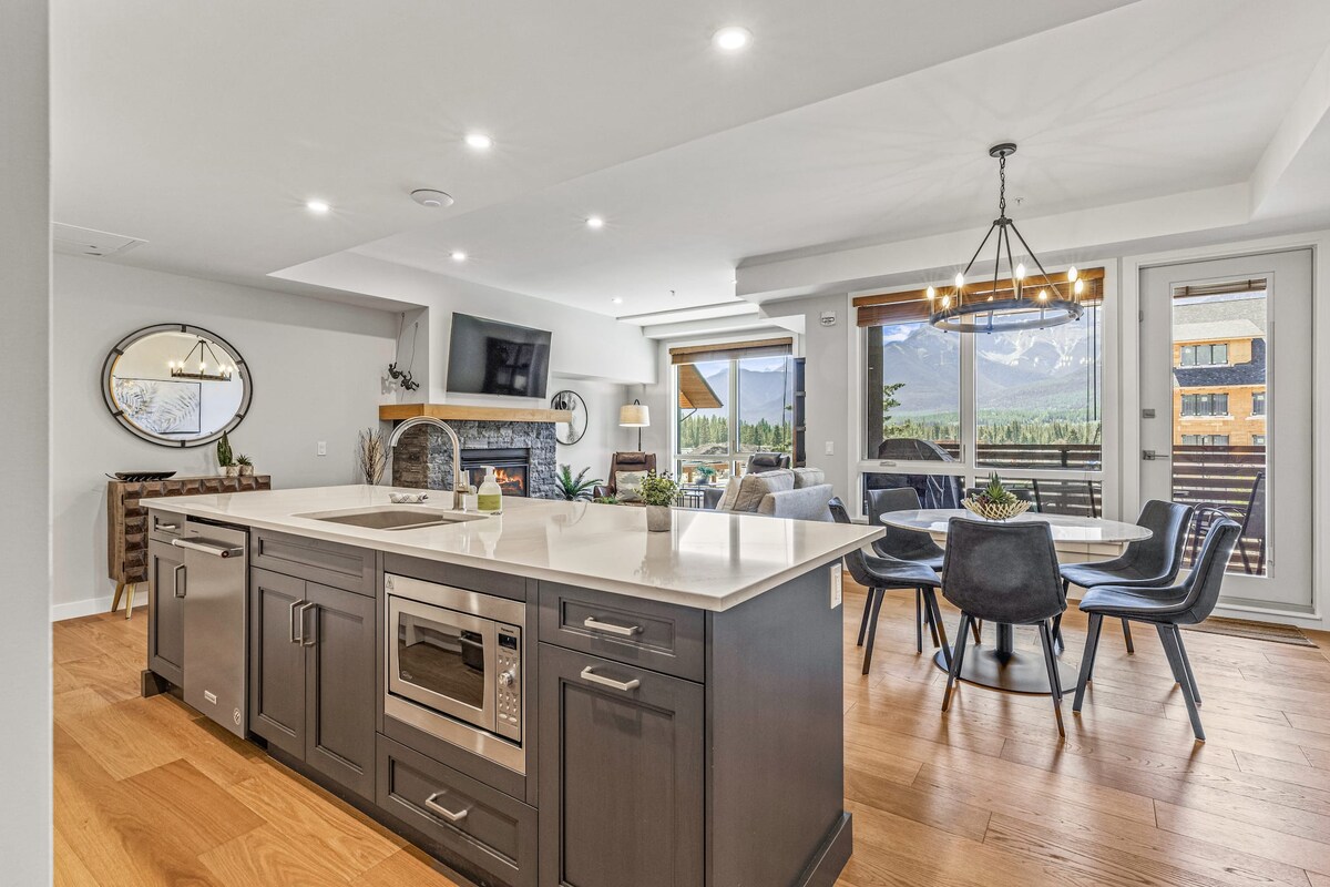 A modern kitchen is highlighted by a large island with sleek countertops and stainless-steel appliances. The open layout connects to a dining area featuring a circular chandelier and a table surrounded by upholstered chairs. Large windows frame scenic mountain views, inviting natural light.