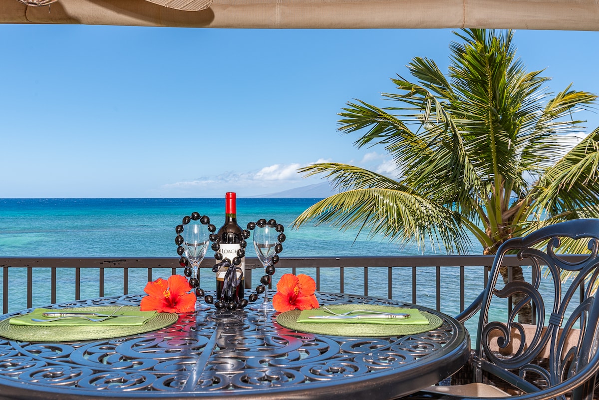A dining table is set on a lanai, featuring two place settings with bright green placemats and glasses. A bottle of wine is placed in the center, flanked by decorative heart-shaped arrangements. Palm trees provide a tropical backdrop against the ocean and blue sky.