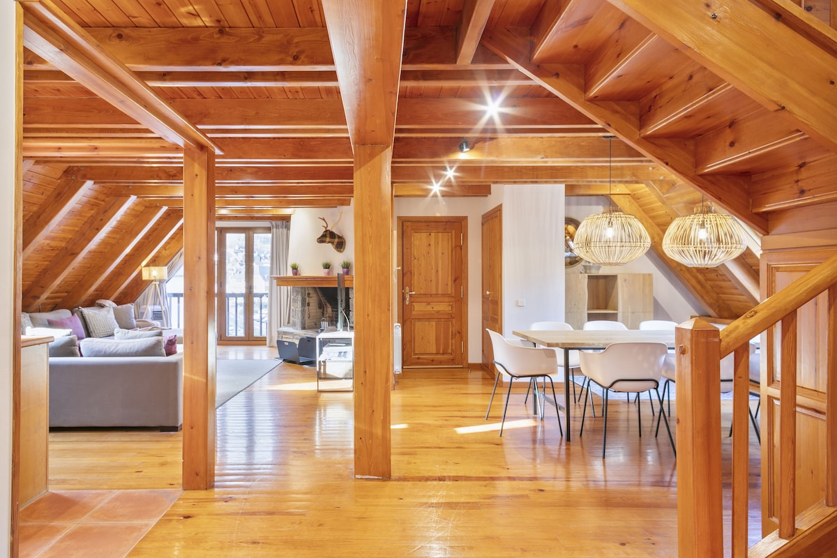 The open-plan living area showcases wooden beams and warm hardwood flooring. A cozy seating arrangement is visible beside a fireplace, while a white dining table with elegant chairs is positioned to welcome gatherings. Natural light filters through large windows, enhancing the inviting ambiance.