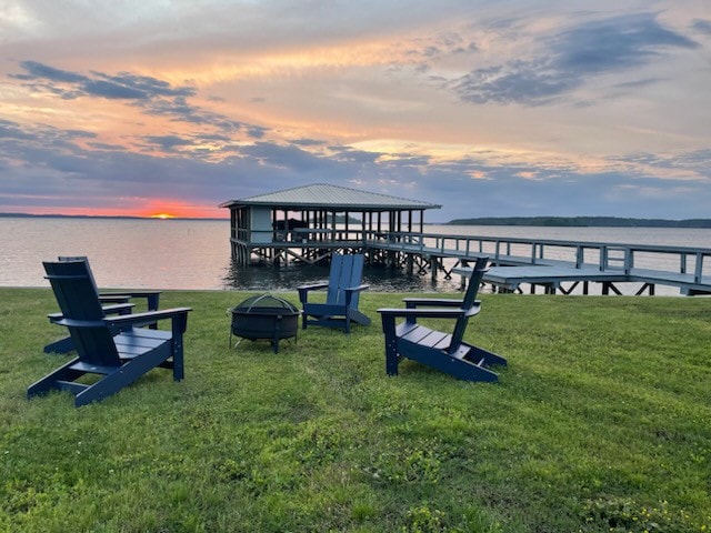 Four blue Adirondack chairs are arranged on a green lawn, facing a serene lake at sunset. A dock extends into the water, featuring a covered structure in the background. The sky displays a blend of warm colors, enhancing the tranquil setting.