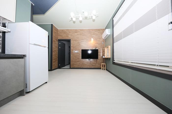 A spacious interior is visible, featuring light-colored flooring and green accent walls. A refrigerator stands on the left, while a television is mounted on the wall ahead. Air conditioning is present, and natural light filters in through the large, partially covered window.