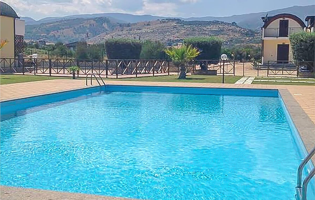 A clear blue swimming pool is seen, with steps leading into the water. Surrounding the pool are grassy areas and landscaped features, including palm trees and lounge chairs. The backdrop features rolling hills under a partly cloudy sky.