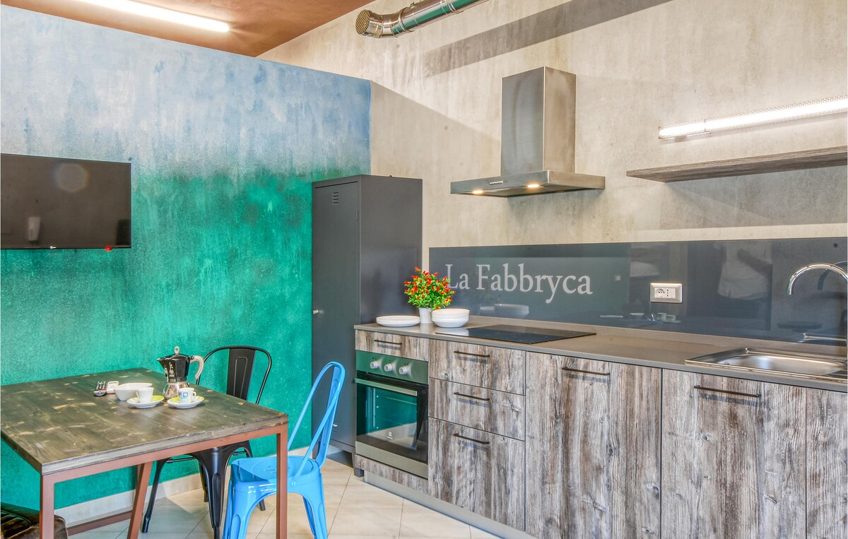 A modern kitchen is highlighted by textured concrete walls in gradient shades of blue and green. Wooden cabinets and a stainless steel oven contribute to the industrial design. A small dining table is set with colorful chairs, complemented by a decorative plant on the counter.