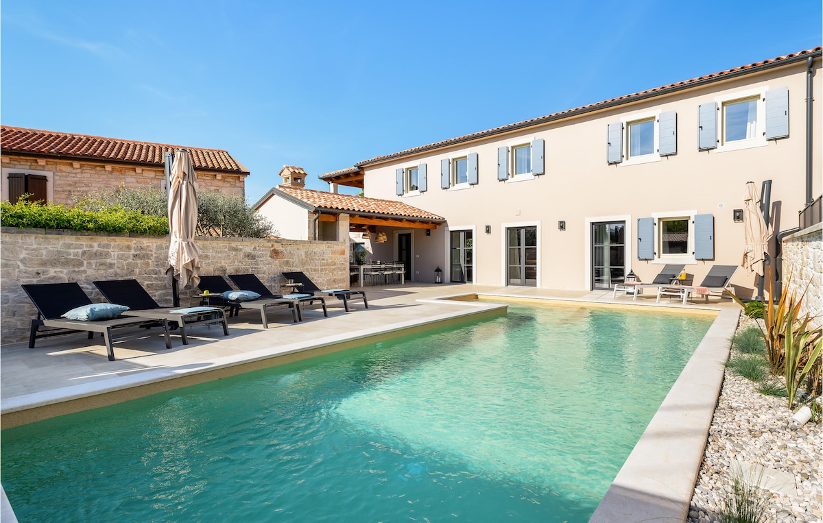 An outdoor area features a crystal-clear private pool bordered by a spacious terrace. Sun loungers are arranged beside the pool, providing ample relaxation space. The two-story villa is visible in the background, showcasing pale exterior walls, blue shutters, and a charming rooftop.