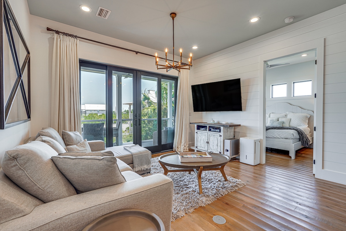 A bright living space is highlighted by large glass doors leading to a covered deck. A comfortable sectional sofa faces a wooden coffee table, with a flat-screen TV mounted on the wall. Natural light fills the room, enhancing the warm wooden flooring.