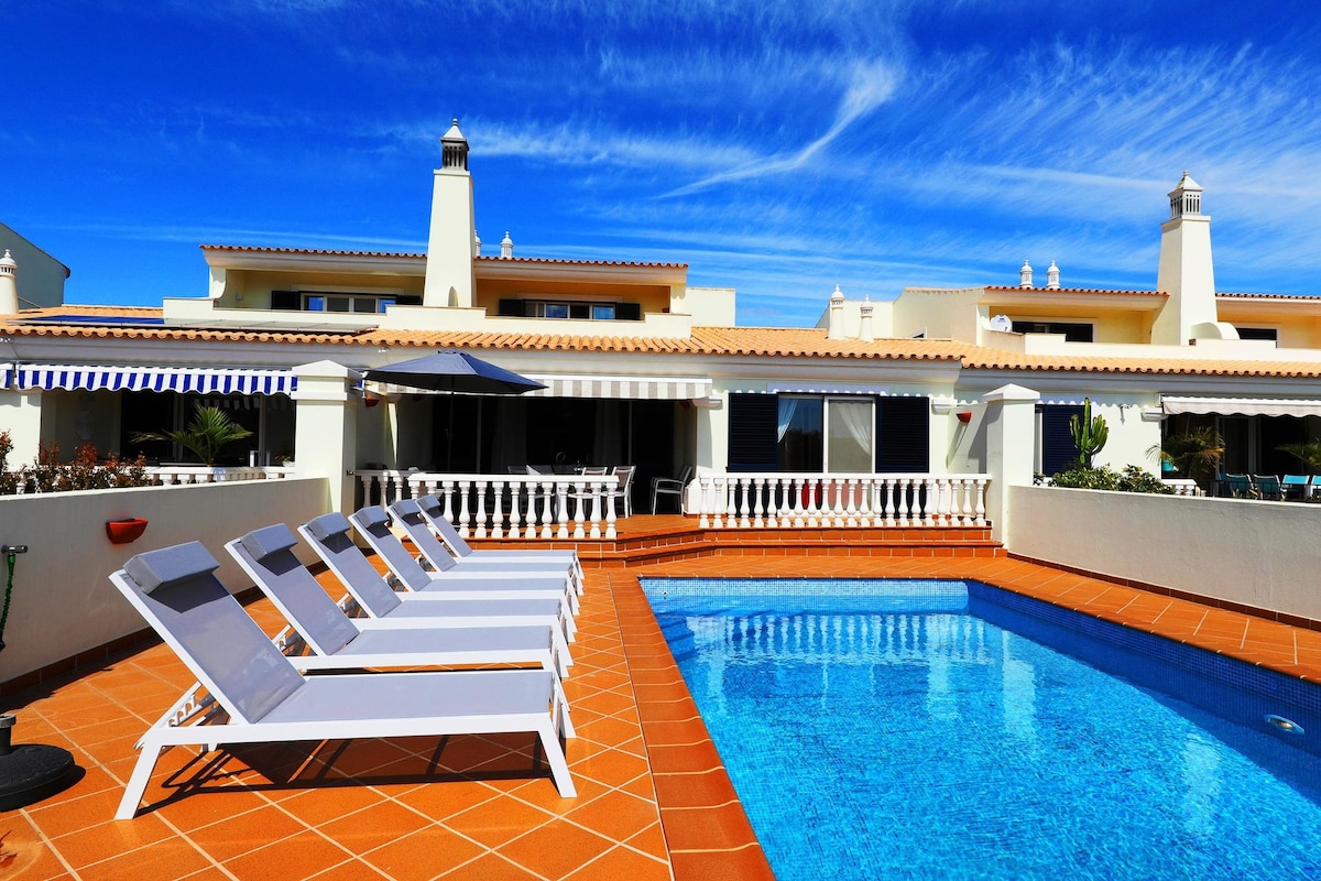A tranquil outdoor space features a sparkling blue pool surrounded by comfortable sunbeds. The sun-filled patio is bordered by white railings, while the villa's spacious exterior displays large windows and bright shades under a clear blue sky.
