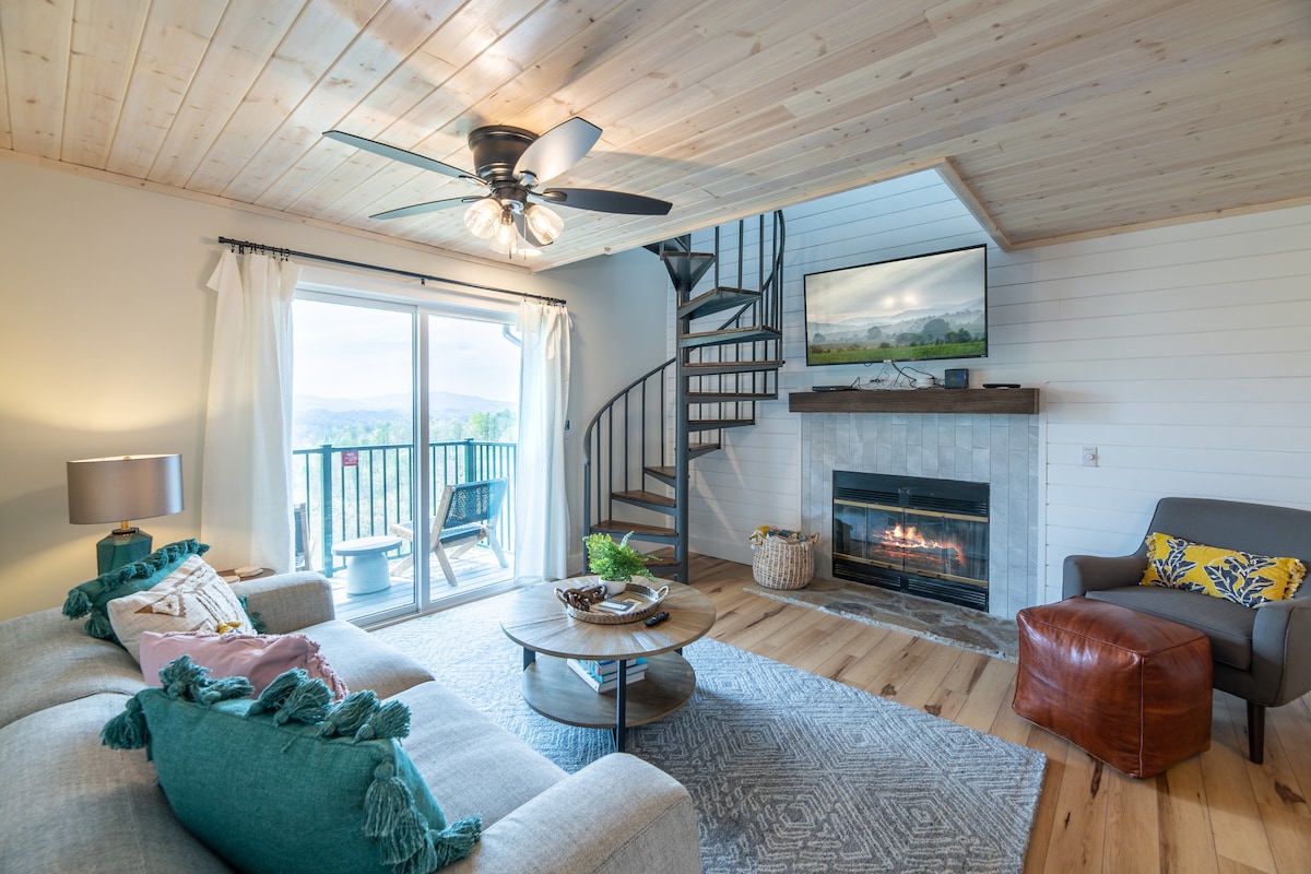 A cozy living area features a spiral staircase leading to the loft bedroom. A wood-burning fireplace is situated beneath a mounted TV, while large glass doors open to a private balcony, allowing natural light to fill the space. Soft furnishings and a round coffee table enhance the inviting atmosphere.