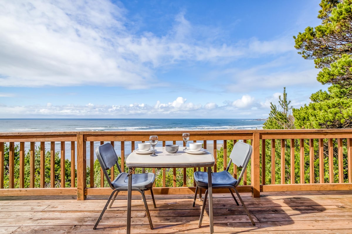 A wooden deck is shown overlooking the ocean, featuring a small table set for two with plates and glasses. The expansive view of the sea is framed by lush trees, and a clear sky filled with scattered clouds enhances the tranquil setting.