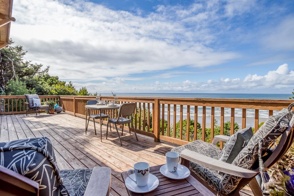 A spacious deck is featured with comfortable seating arrangements and a small dining table, overlooking the ocean. Two cups are placed on a table, with the expansive sea visible in the background, complemented by a clear blue sky and scattered clouds.