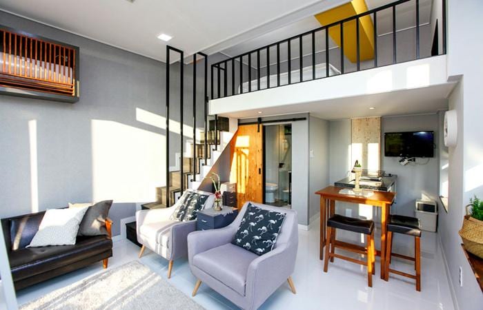 A modern open-concept living area is featured, showcasing a mix of comfortable seating with a dark sofa and light-toned chairs. A dining area with wooden stools is visible, alongside a staircase leading to the upper level, where additional sleeping quarters are situated.