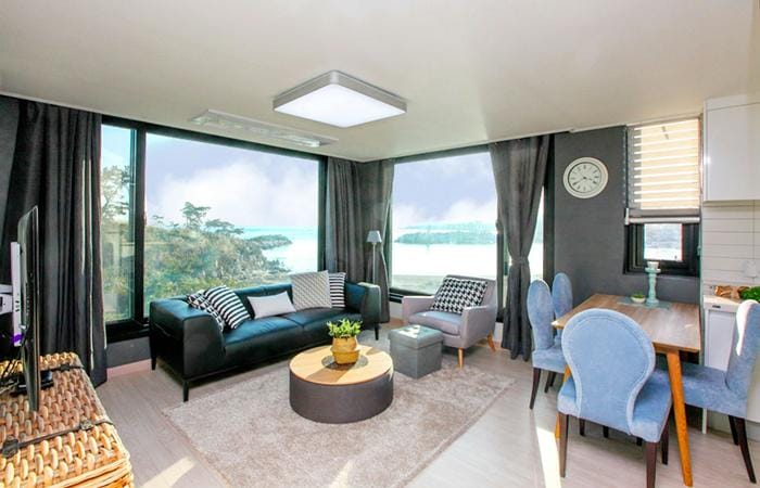 A bright living space features large windows showcasing a coastal view. A dark sofa and two blue chairs surround a circular coffee table, complemented by a light-colored rug. A small dining area is positioned near the kitchen, with a clock adorning the wall.