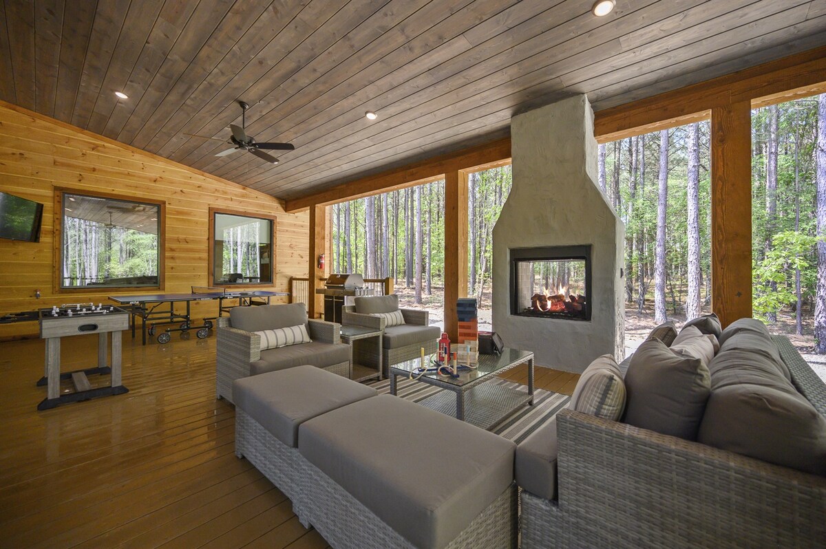 An expansive outdoor living area features a cozy seating arrangement with inviting gray cushions and a glass coffee table. A modern fireplace centers the space, surrounded by floor-to-ceiling windows that showcase the natural beauty of the surrounding pine trees.
