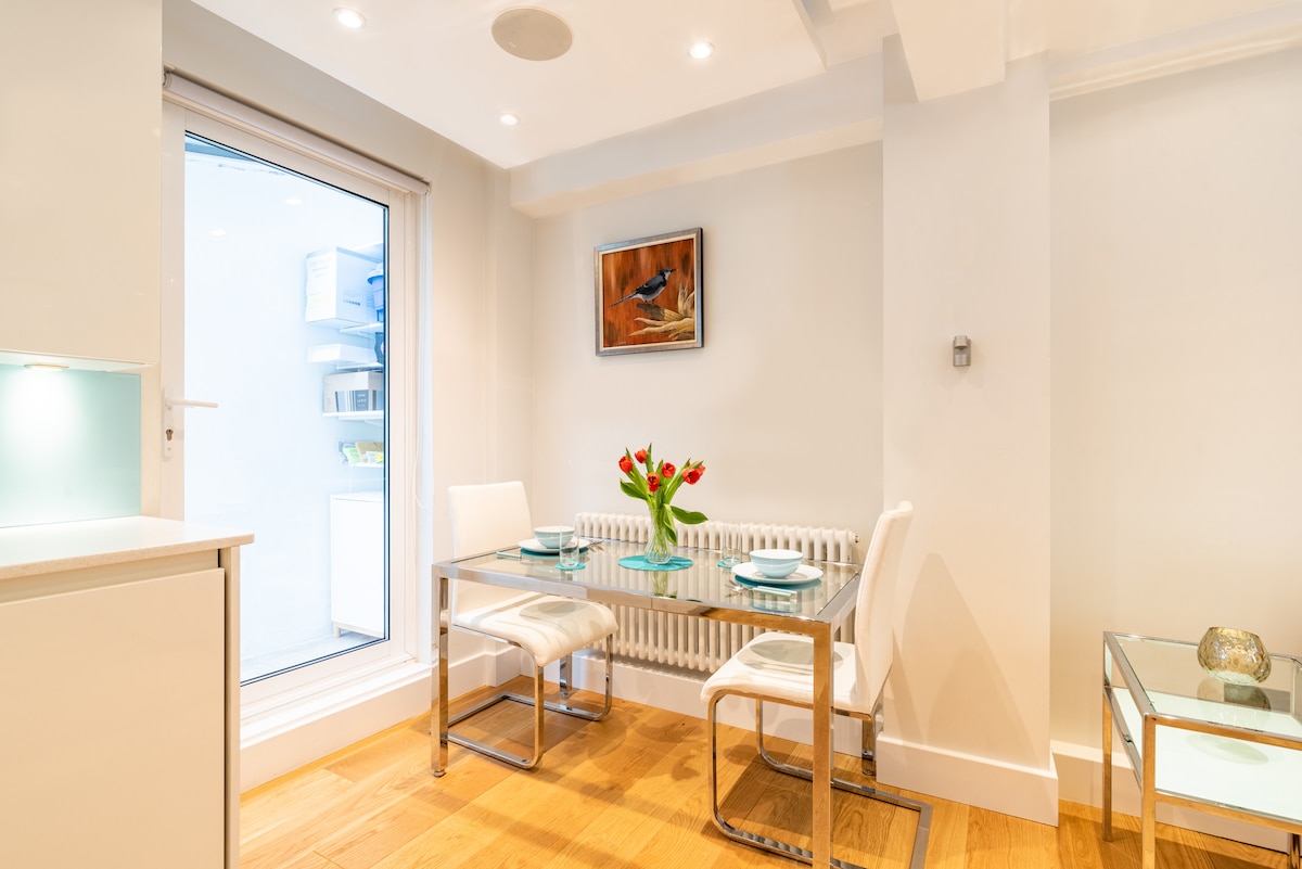 A light-filled dining area features a modern glass table with two white chairs, set with a vase of fresh red tulips. A sliding glass door opens to a small outside space, while soft lighting enhances the bright, minimalist decor.