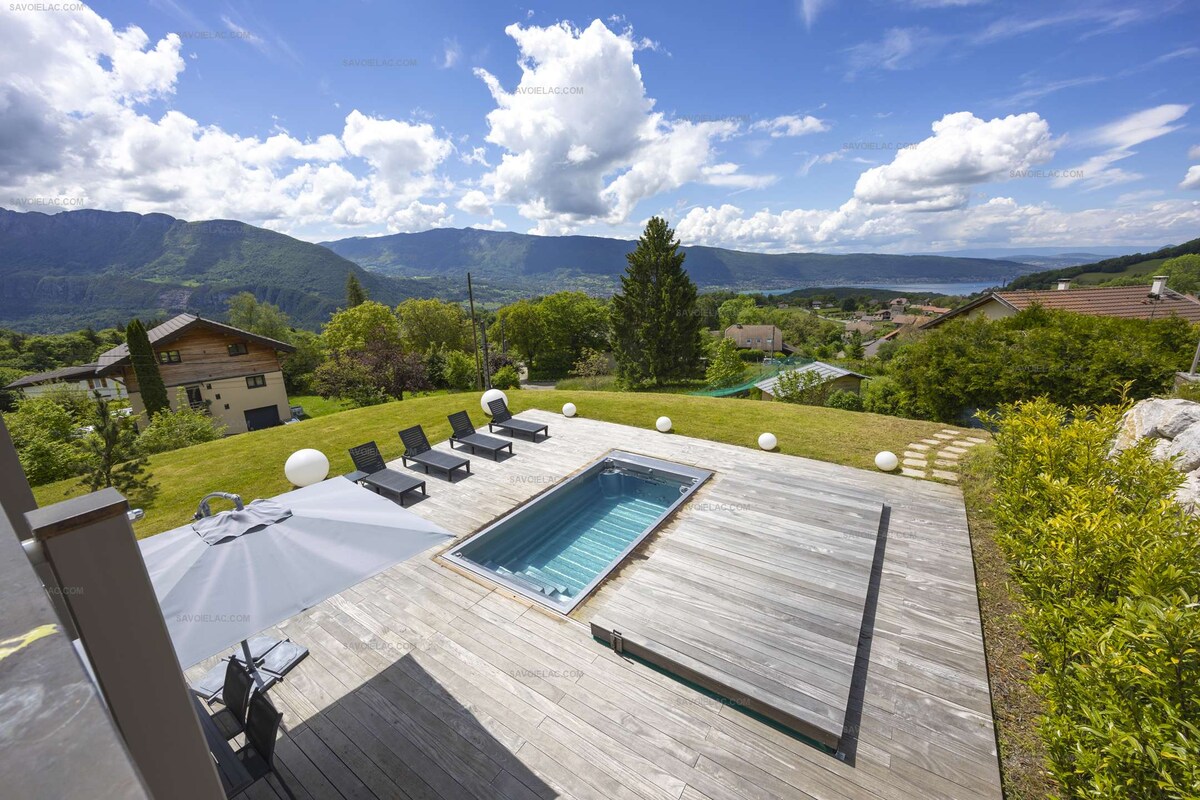 The outdoor space features a heated spa pool set within a wooden terrace. Several sun loungers are arranged around the pool, while the surrounding area is complemented by green lawns and landscaped gardens. Scenic views of the mountains and lake are visible in the background.