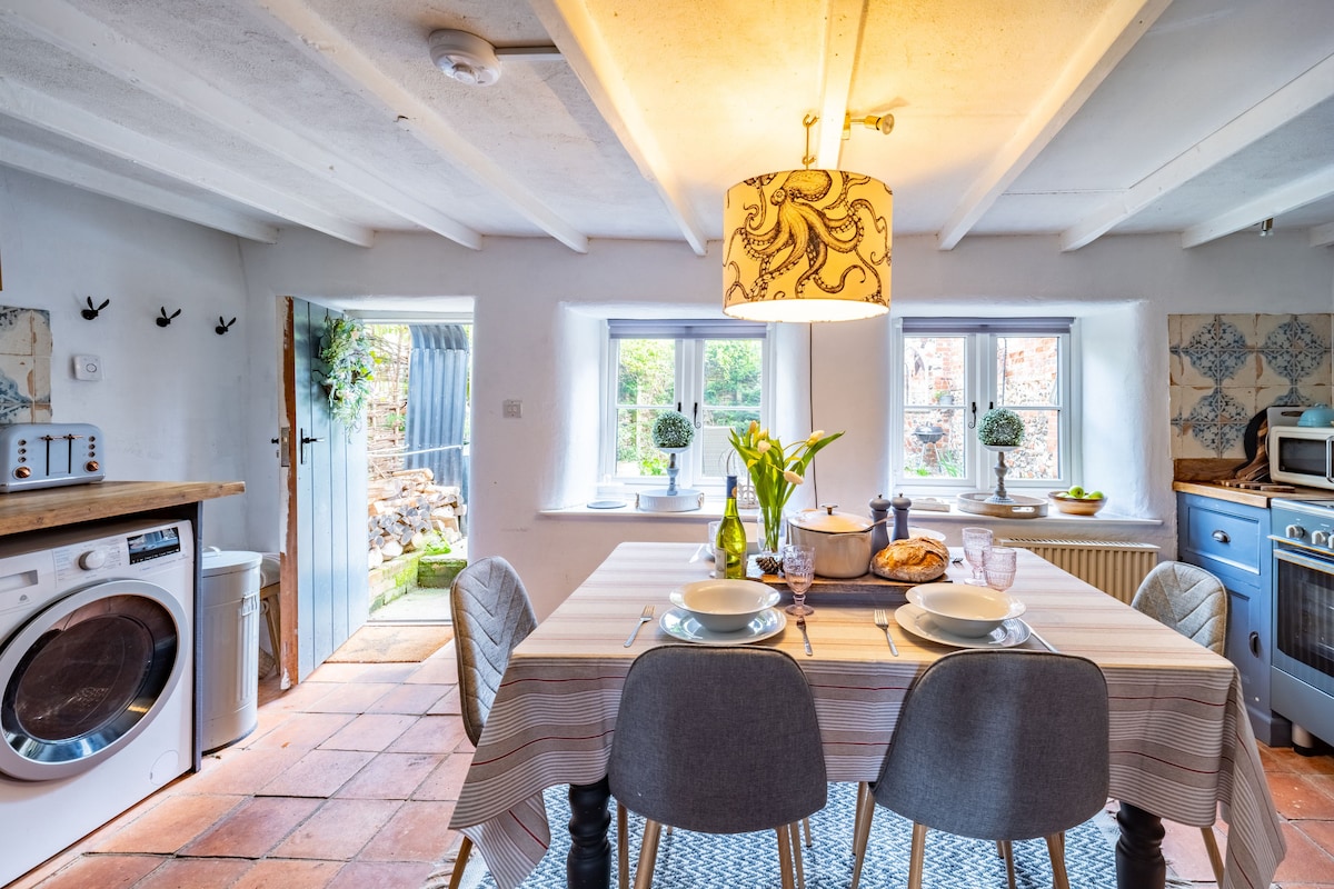 A charming kitchen/diner is featured with a rustic table set for four, complete with dishes and a vase of fresh flowers. Modern appliances, including a washing machine and electric cooker, complement traditional Portuguese wall tiles and light-filled windows that overlook a garden.