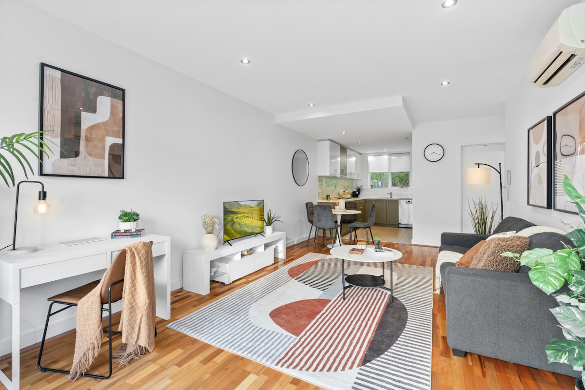 An open plan living area is highlighted by a vibrant area rug, surrounded by a comfortable sofa and chair arrangement. A dining table for four is visible, with a well-equipped designer kitchen in the background. Natural light brightens the space, enhanced by minimal decor.