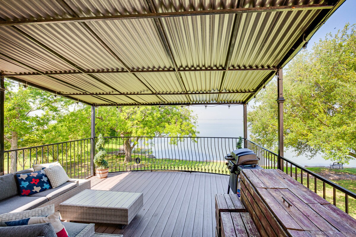 A covered outdoor deck features lounge furniture and a dining setup, ideal for gatherings. A gas grill is positioned nearby, with views of the lake visible beyond a decorative railing. Lush greenery surrounds the area, enhancing the peaceful environment.